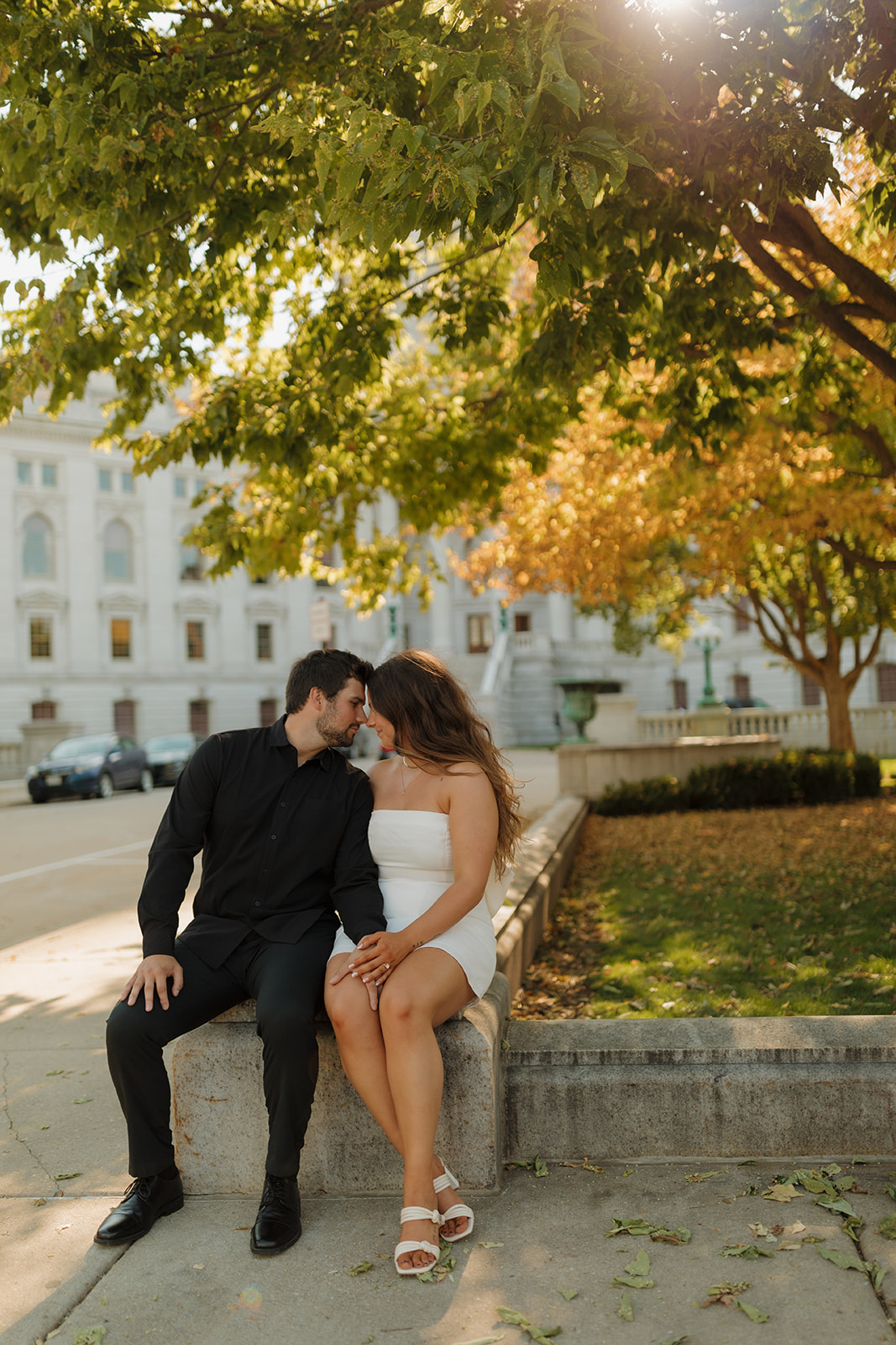 Outdoor engagement photos of a couple sitting on a stone ledge under golden fall leaves near the Wisconsin State Capitol.