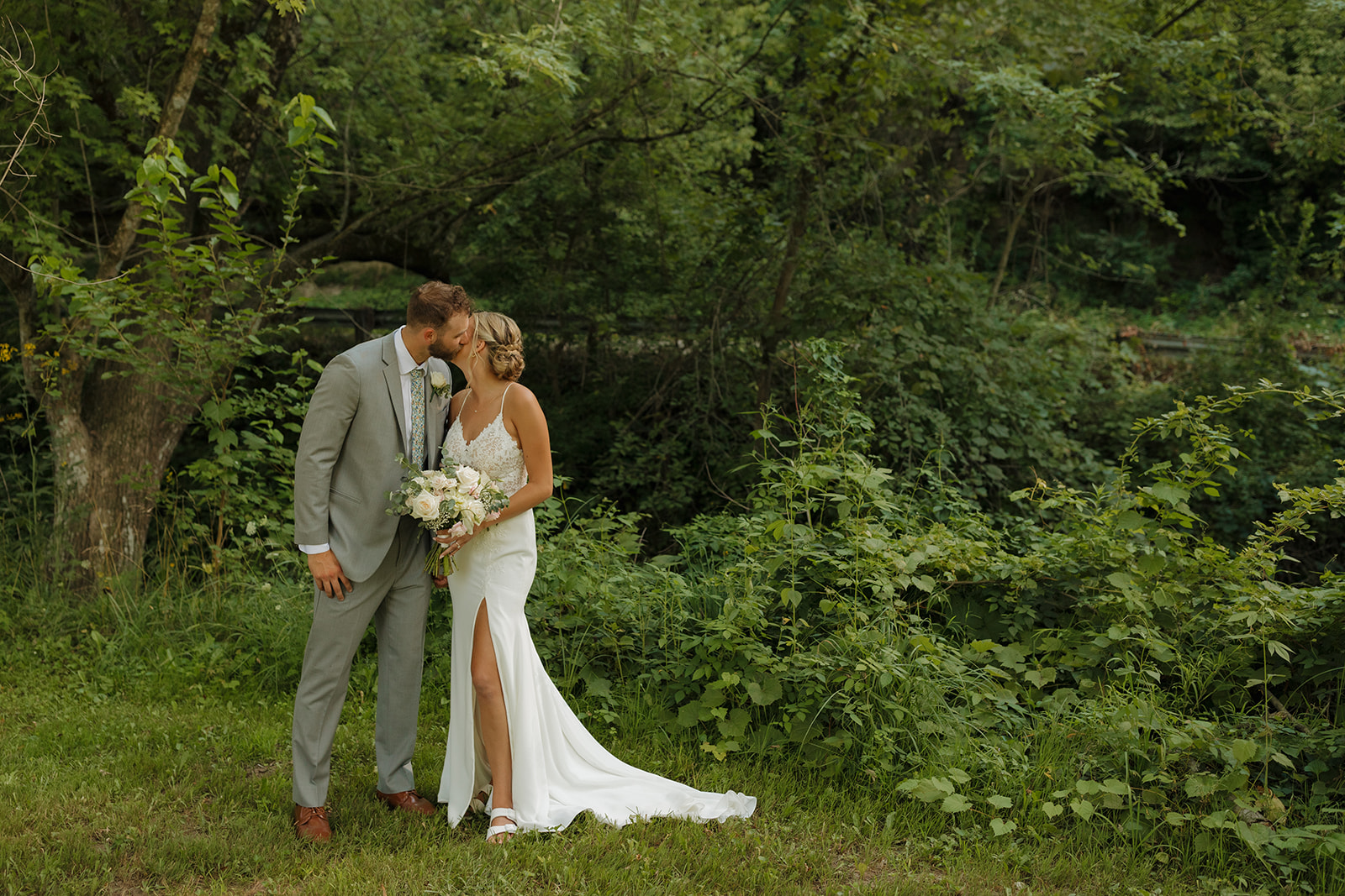Groom kisses bride on cheek in front of tall grassy meadow during bride and groom photos