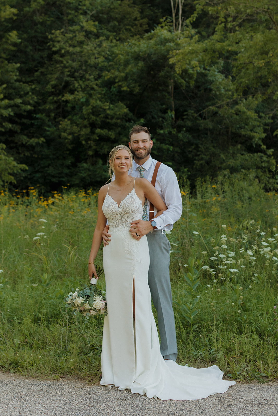 Groom in white button down shirt and suspenders stands with bride in lace wedding gown in front of grassy meadow