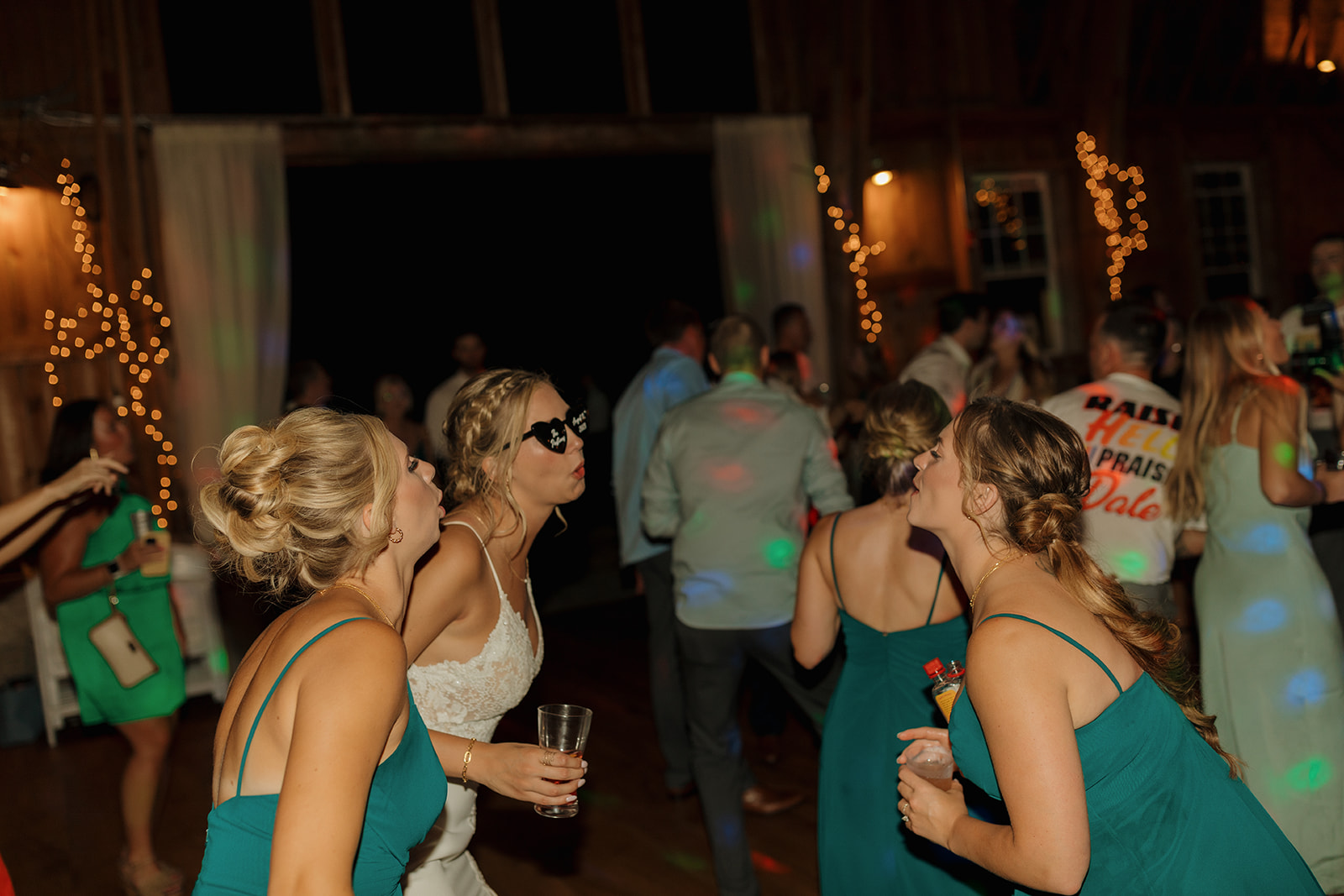 Bride dancing with her bridesmaids on dance floor wearing black heart sunglasses and making silly faces
