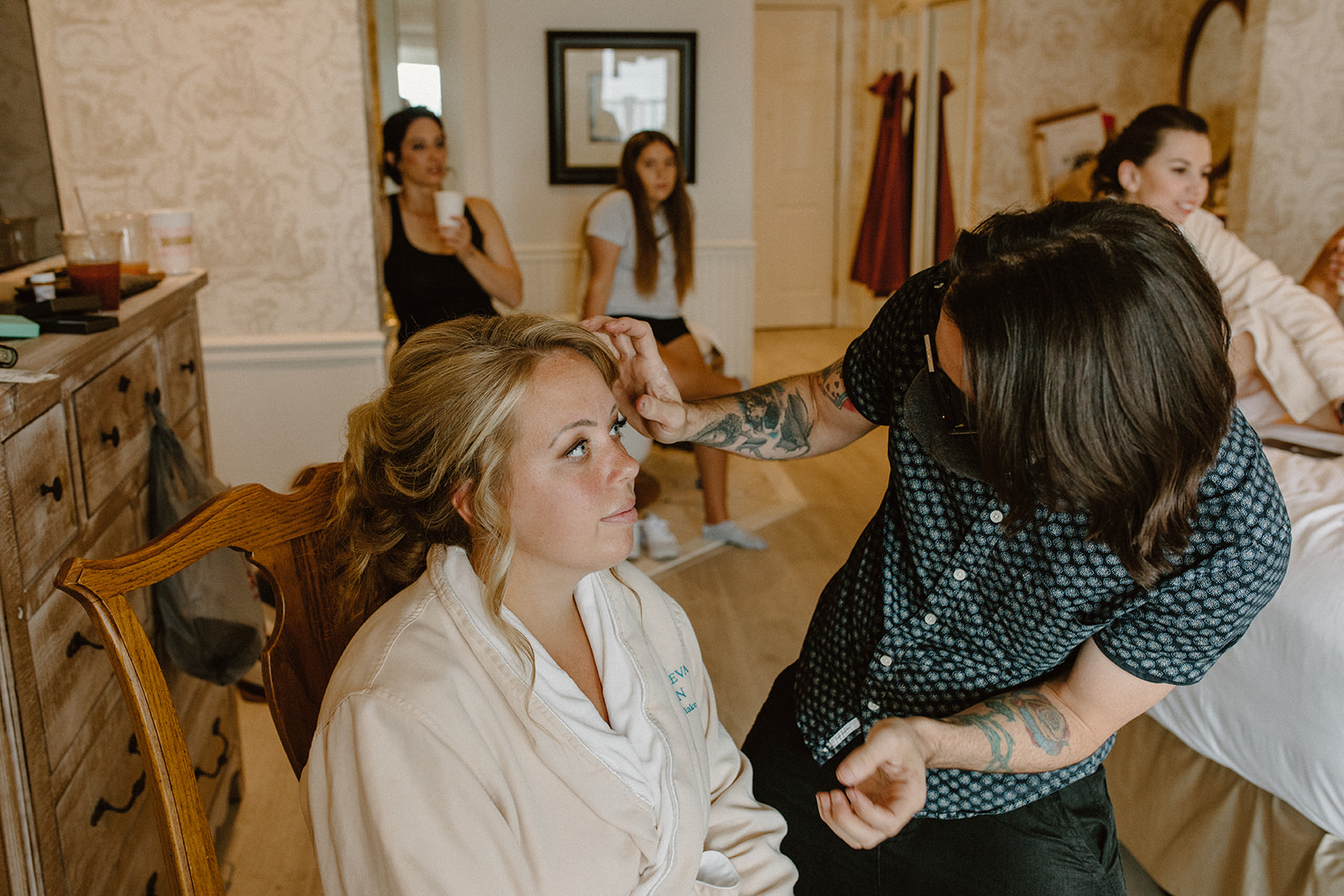 Bride smiling up at makeup artist during wedding morning prep with bridesmaids in the background