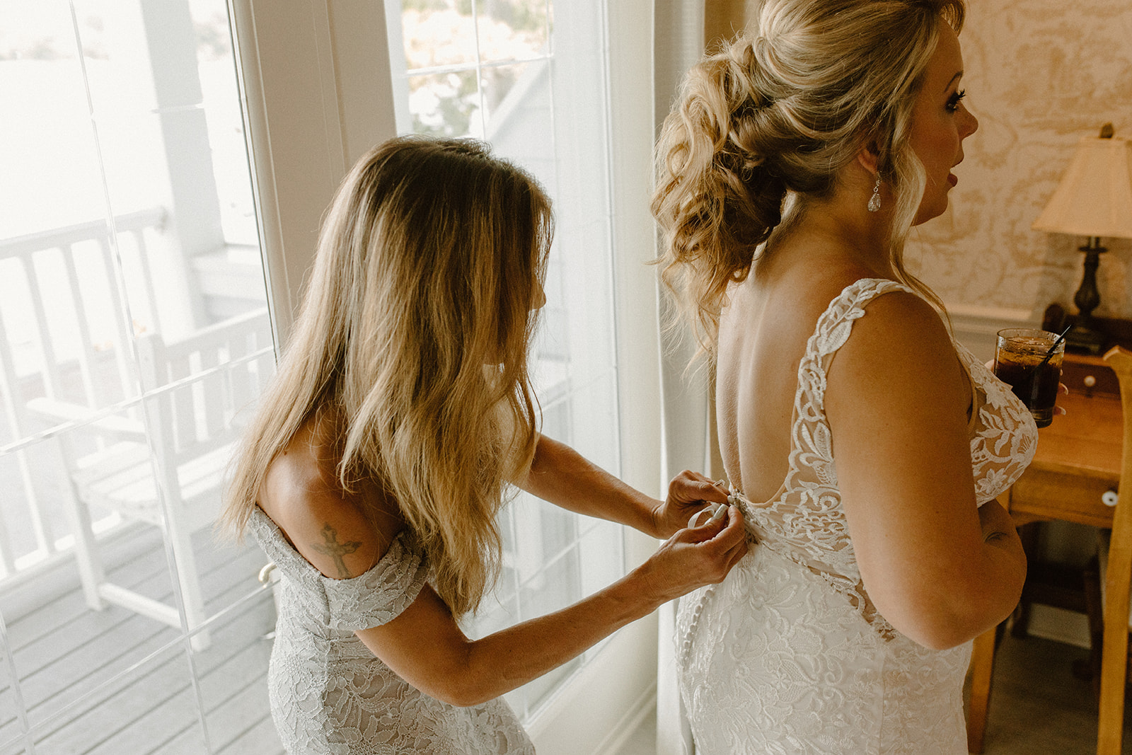 Bride getting zipped into her wedding gown by bridesmaid near a sunlit window