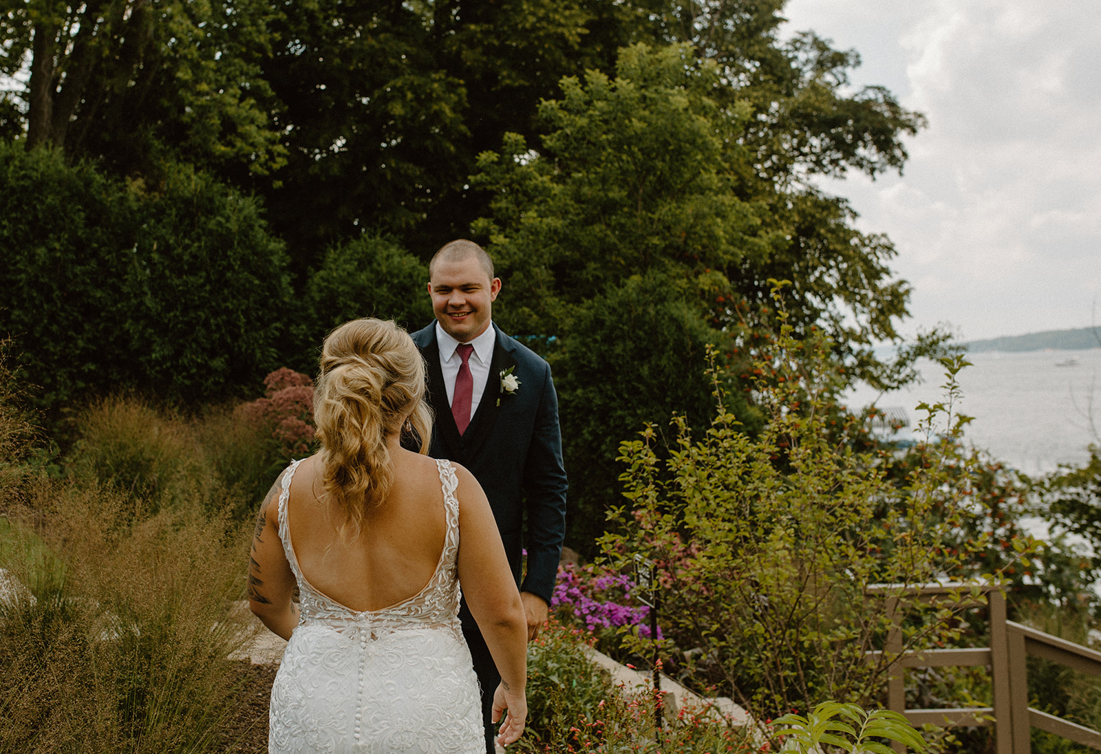 First look moment with groom beaming as the bride walks toward him through lush garden landscaping