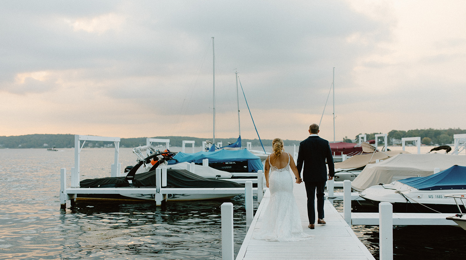 Newlyweds walking hand-in-hand down a white dock surrounded by boats and water during their Lake Geneva wedding
