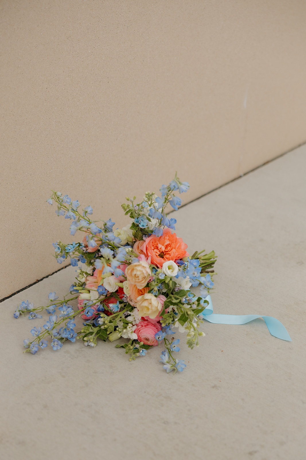 Colorful spring wedding bouquet with peach, pink, and blue blooms and a light blue ribbon.