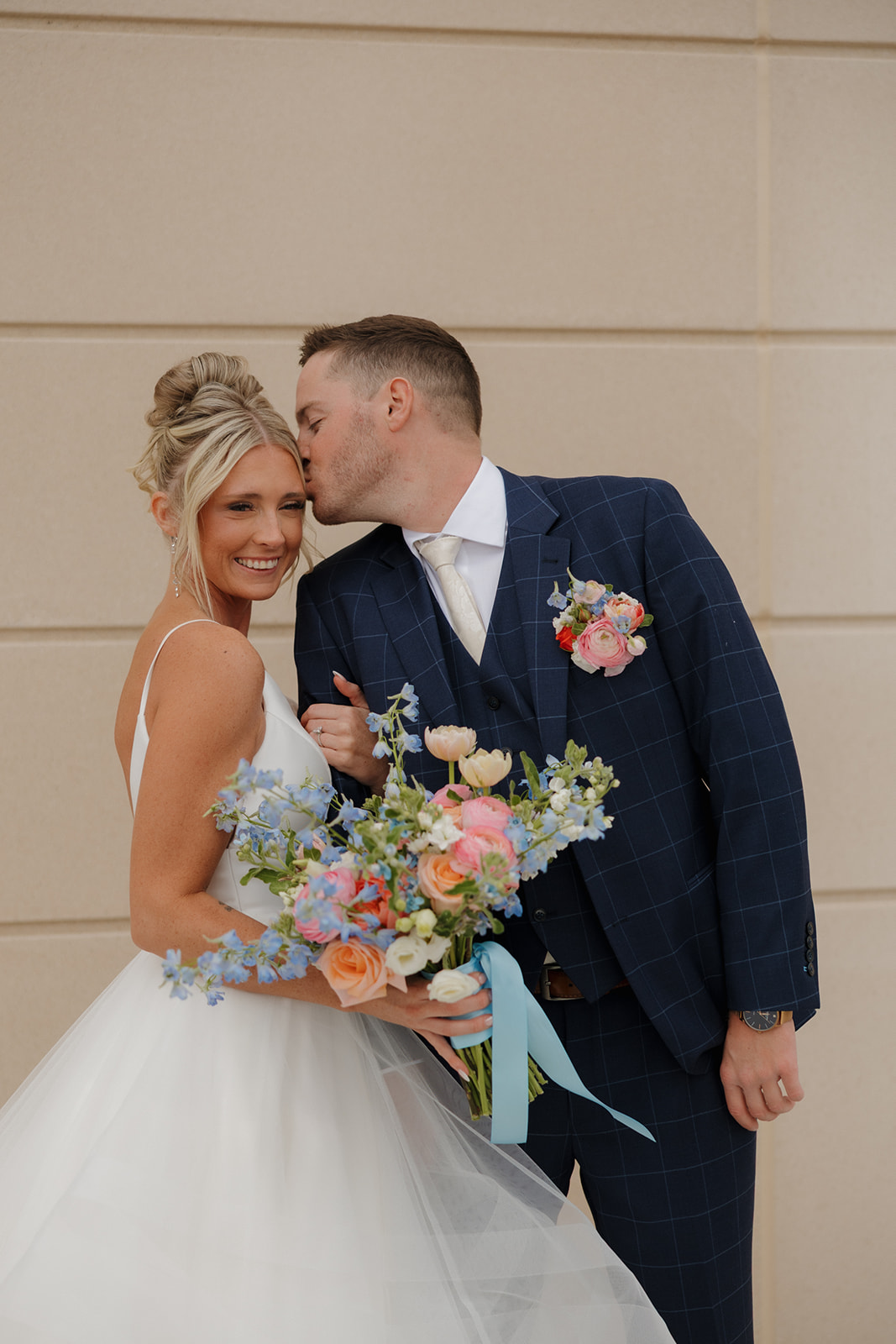 Groom kisses his smiling bride on the temple as she holds a colorful spring bouquet—pure Madison Wisconsin wedding bliss.
