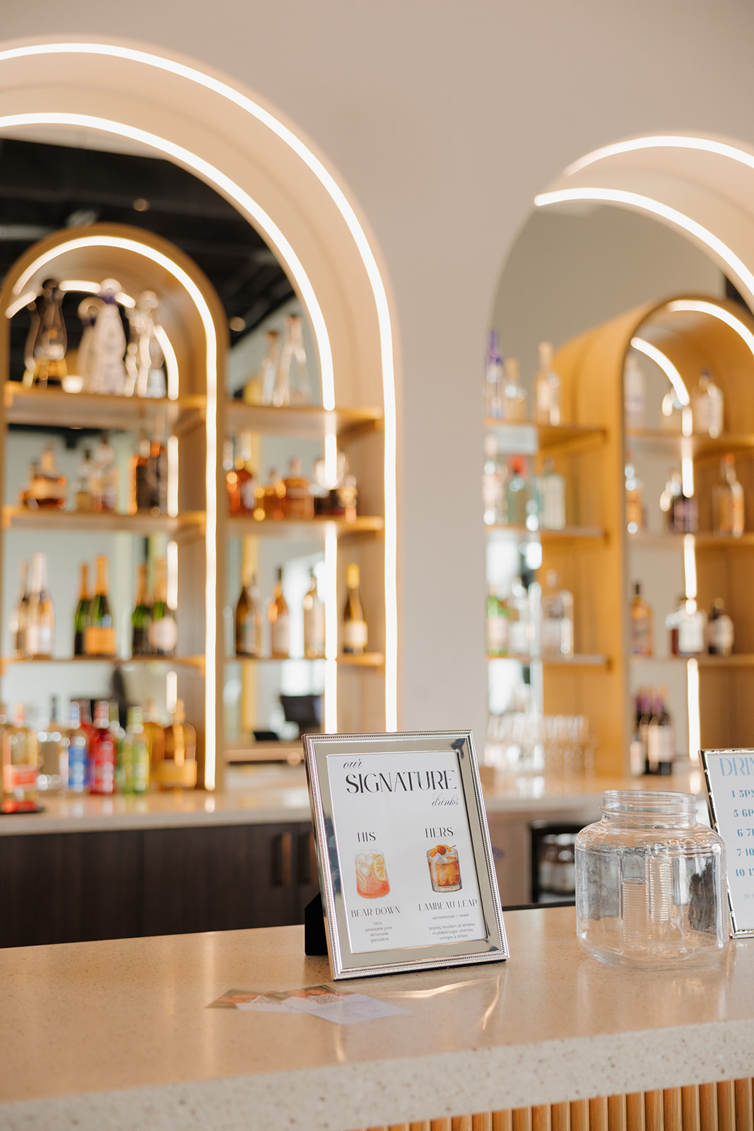 Modern wedding bar setup with backlit shelves and framed signature drink menu.

