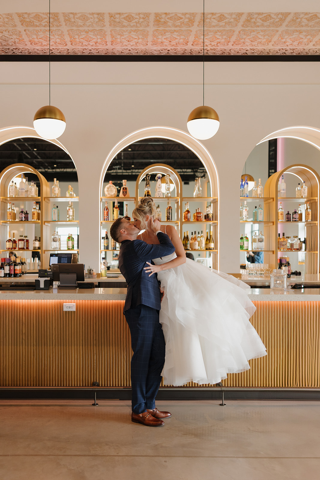 Groom lifts bride for a joyful kiss in front of an art deco-style bar with golden arches and glowing shelves.