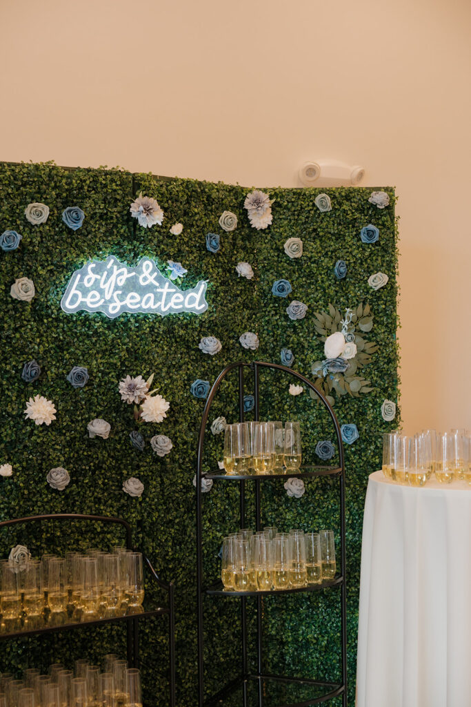 Full view of greenery champagne wall with white and blue florals and elegant flute displays at a spring wedding. 