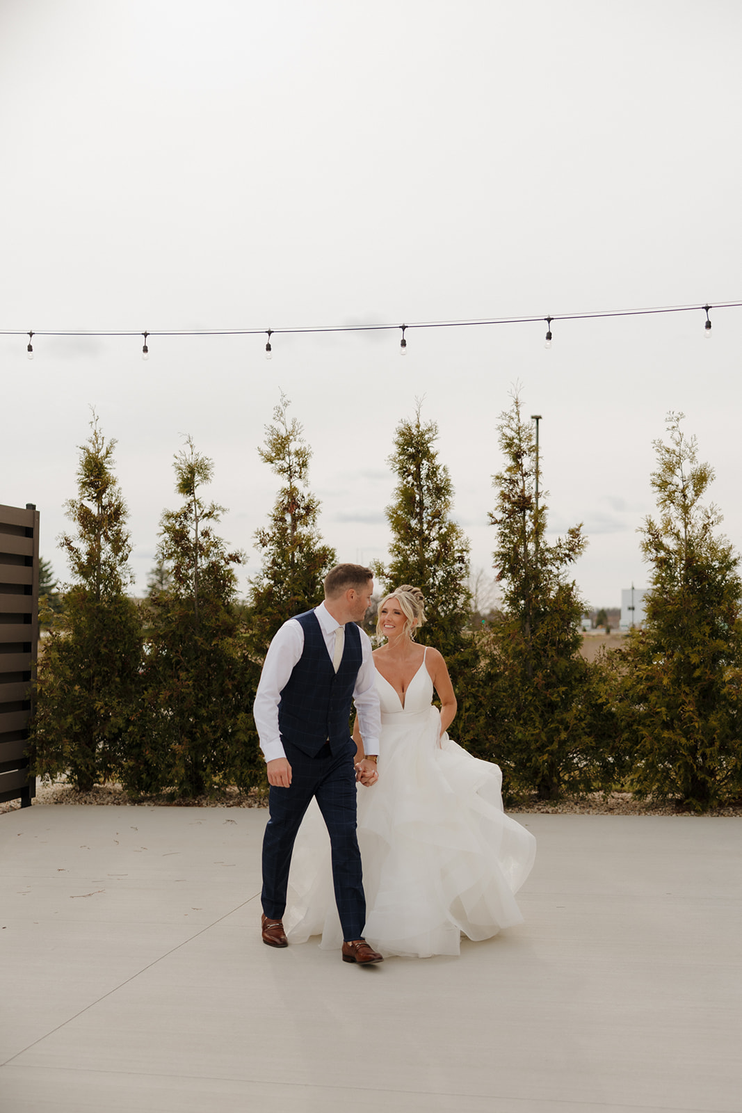Bride and groom walk hand-in-hand under string lights with trees lining the background.