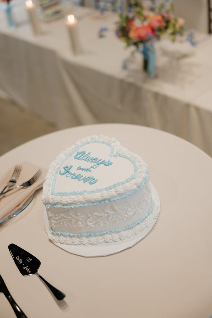 Heart-shaped wedding cake with “Always and Forever” piped in blue, styled on a sweetheart table.

