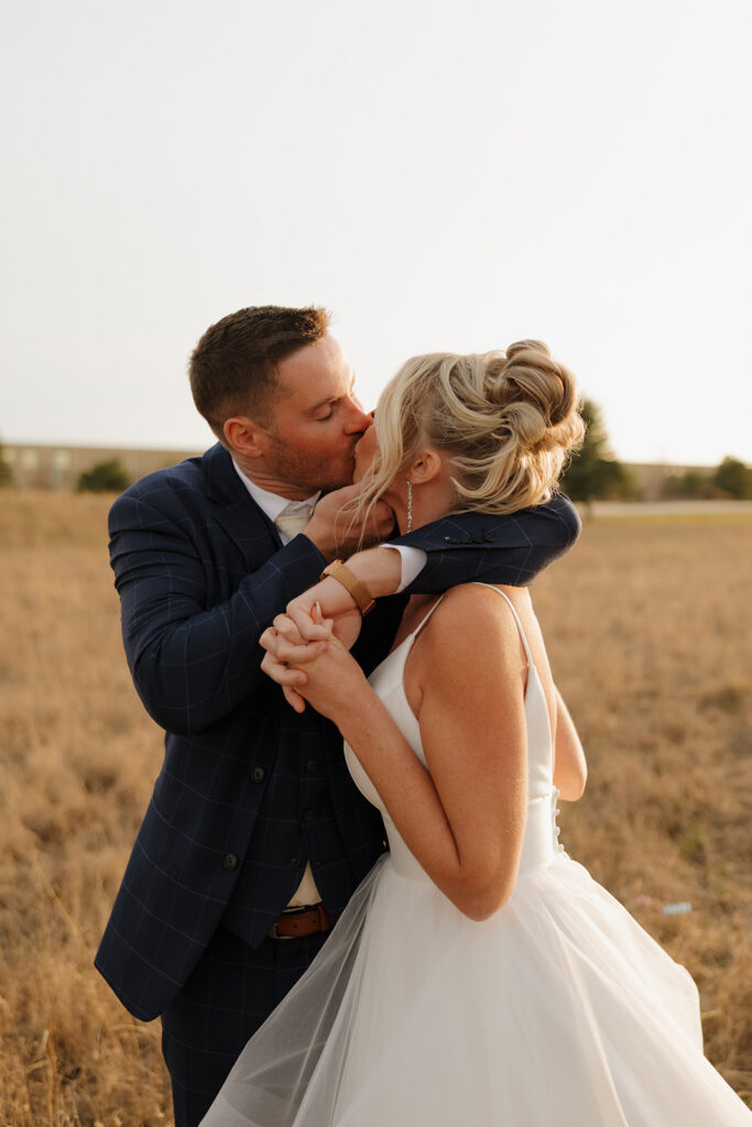 Groom pulls bride in for a passionate kiss with their hands intertwined during golden hour at their Madison Wisconsin wedding