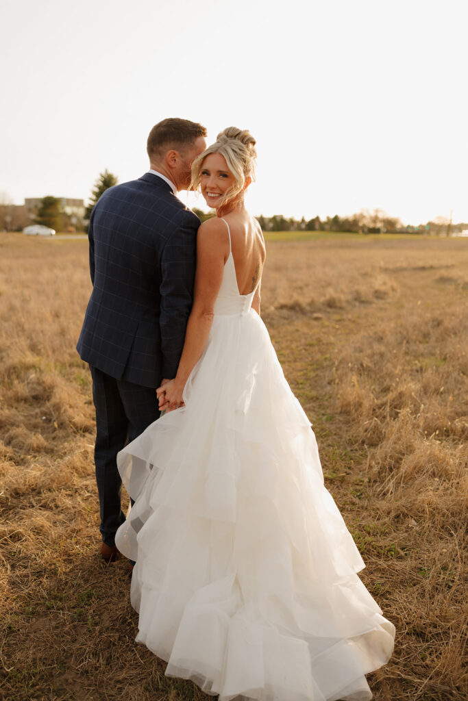 Bride smiles back at the camera while holding her groom’s hand, dress trailing behind her in a golden meadow. 
