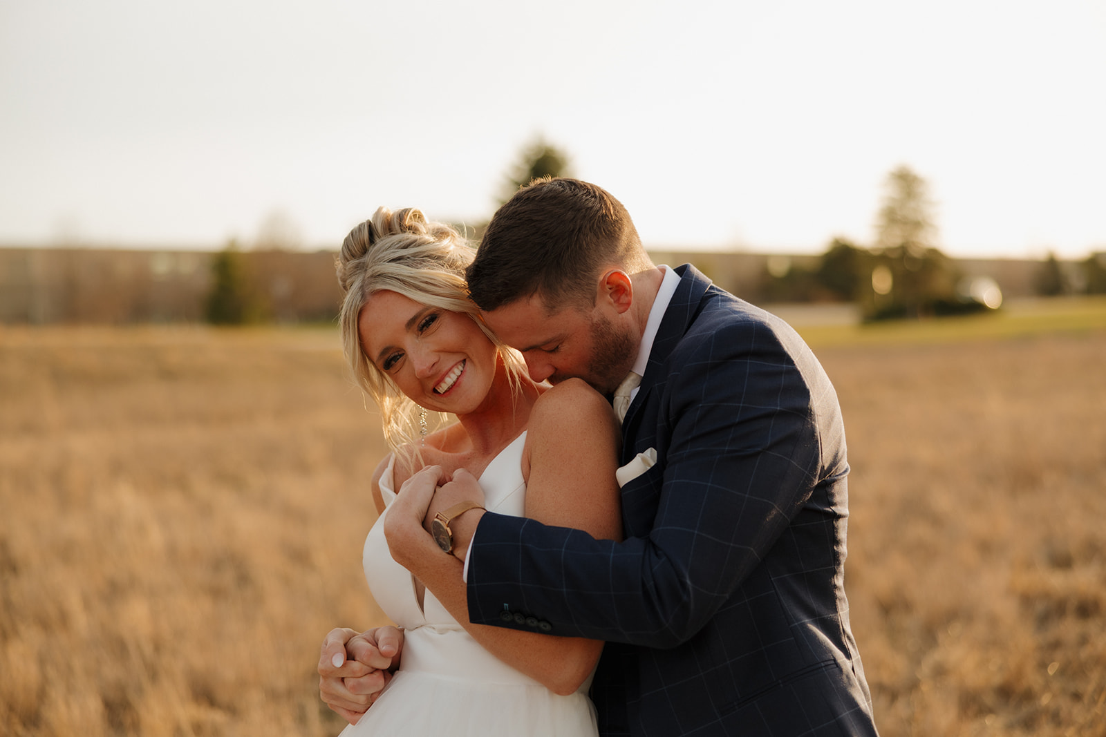 Groom kisses bride’s shoulder during golden hour in a romantic field outside Madison, Wisconsin.