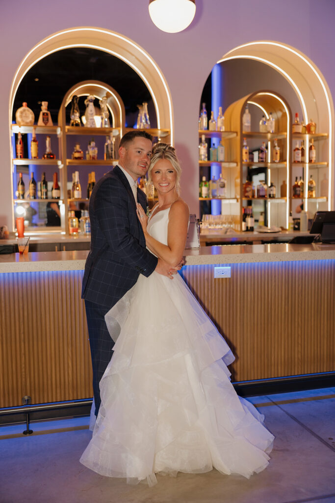 Bride and groom pose in front of a modern, glowing wedding bar with curved arches and warm lighting.