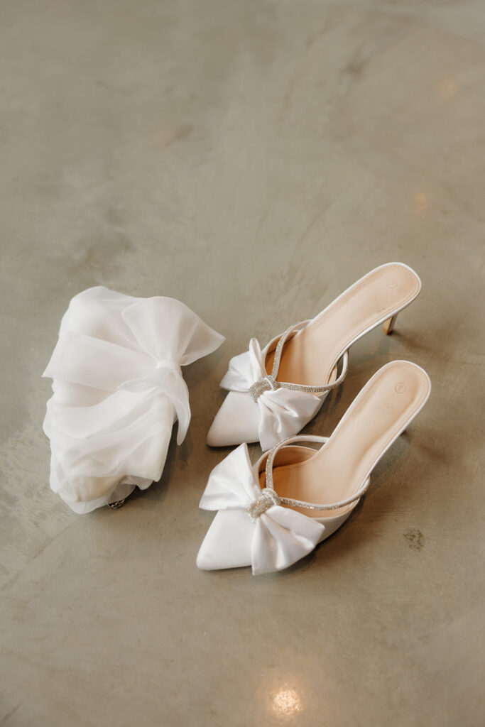 Elegant bridal accessories laid out on concrete: white bow heels and a delicate organza clutch.