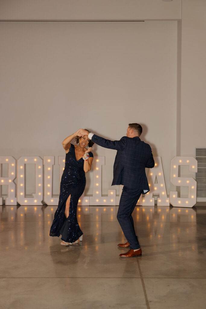 Groom twirls his mom during their dance, surrounded by soft lighting and marquee letters spelling their name