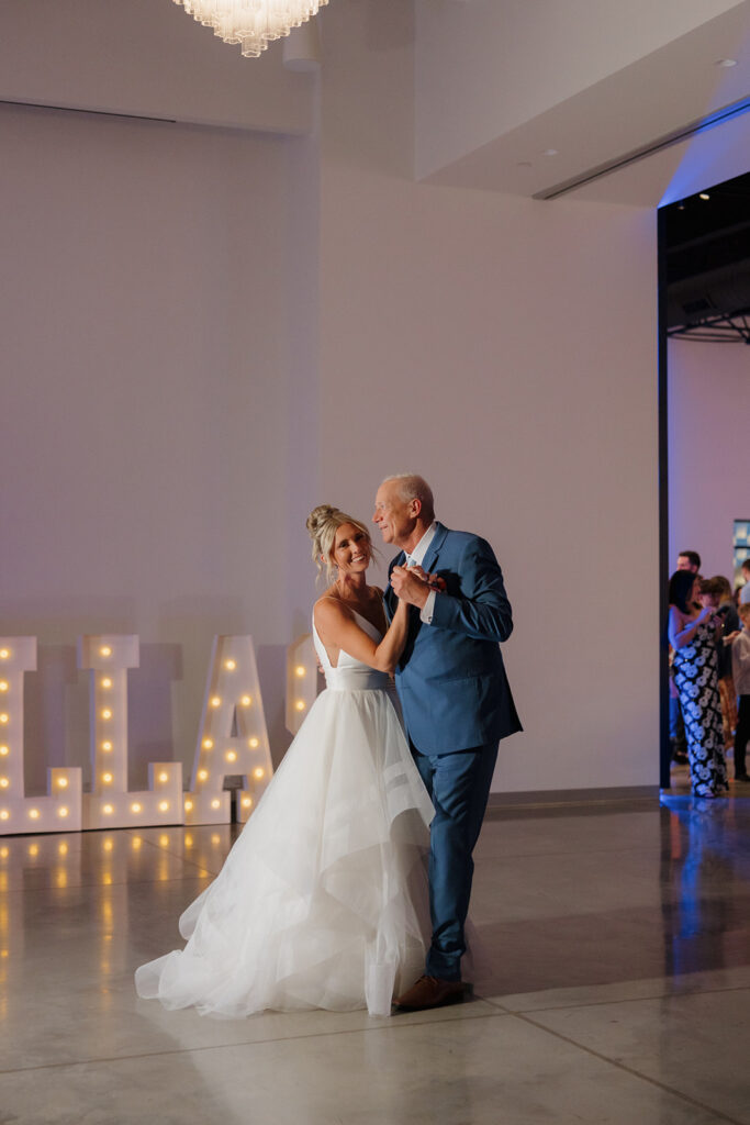 Bride shares an emotional father-daughter dance under a chandelier, smiling sweetly in her dad’s arms.