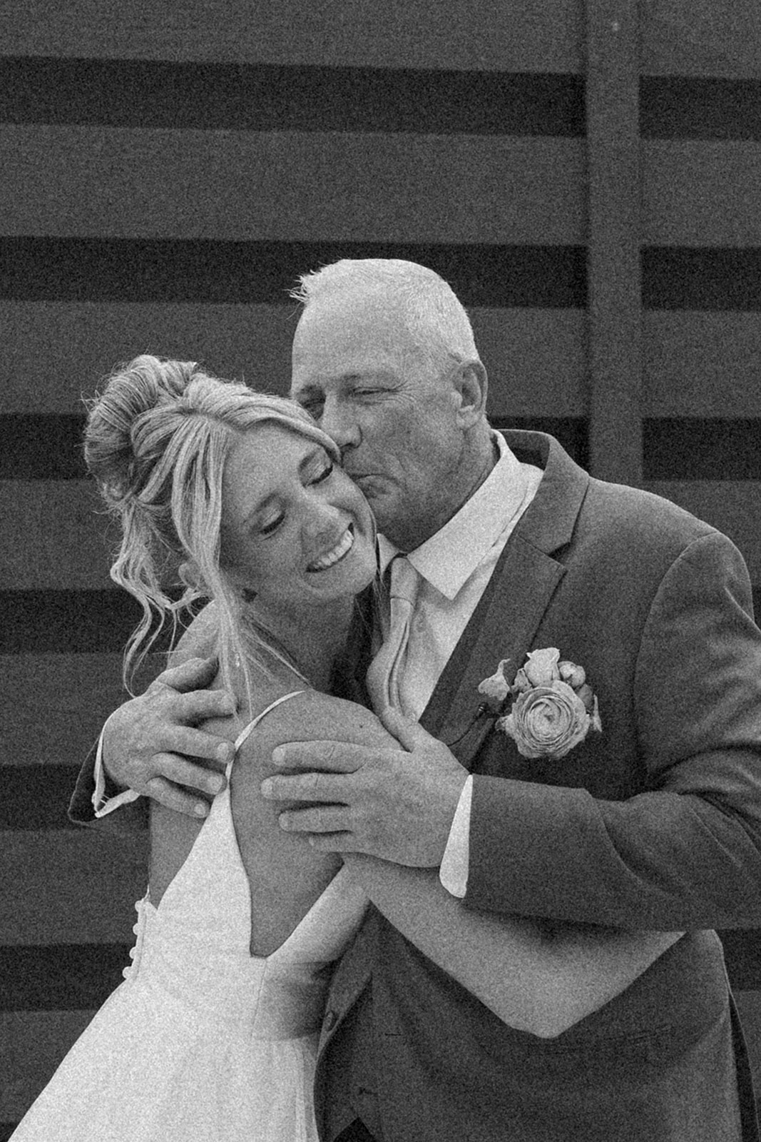Emotional black and white photo of a bride hugging her dad while he kisses her cheek