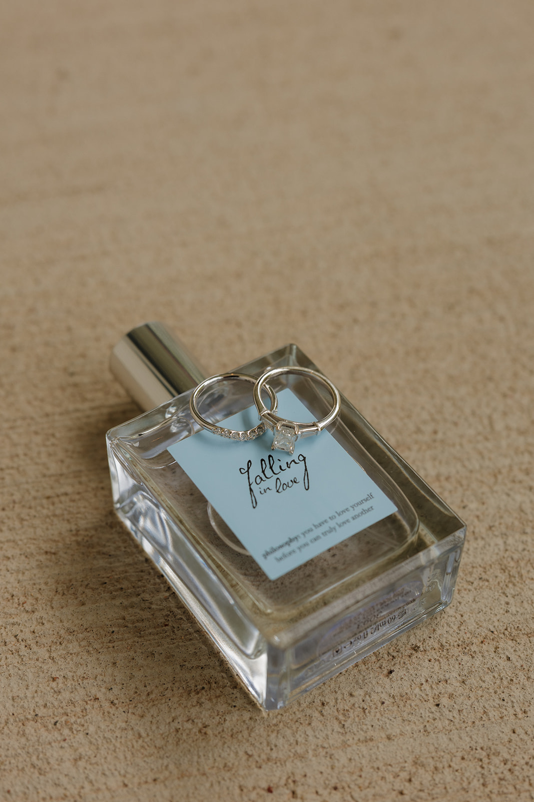 Close-up of wedding rings resting on a bottle of “Falling in Love” perfume—captured as part of elegant country club wedding details.