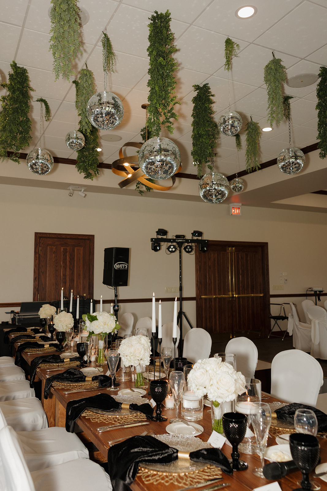 Elegant country club wedding reception setup with black and gold accents, white floral centerpieces, taper candles, and disco balls suspended above the tables.