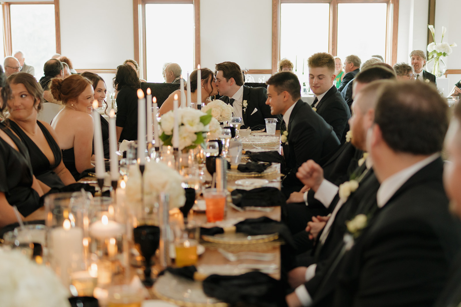 Bride and groom share a kiss at the head table during their country club wedding reception, surrounded by the wedding party and candlelit florals.