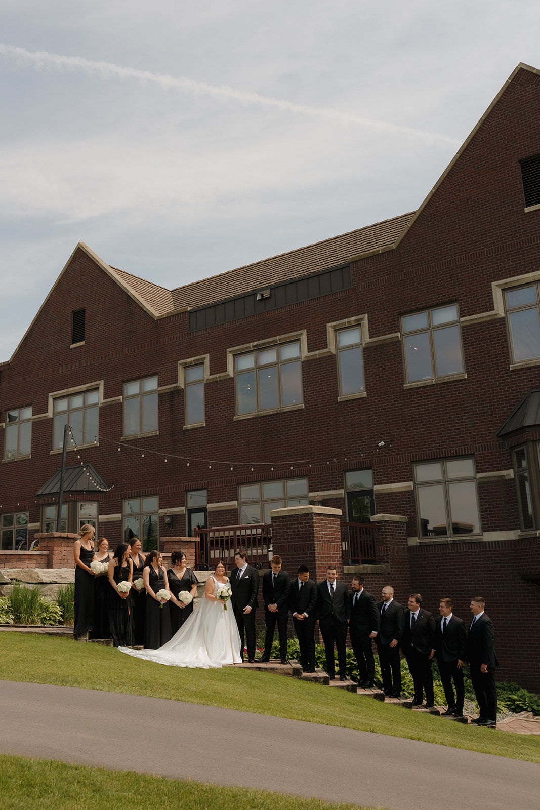 Bride and groom with their full wedding party lined up outside a brick country club wedding venue, dressed in black formalwear with white bouquets.