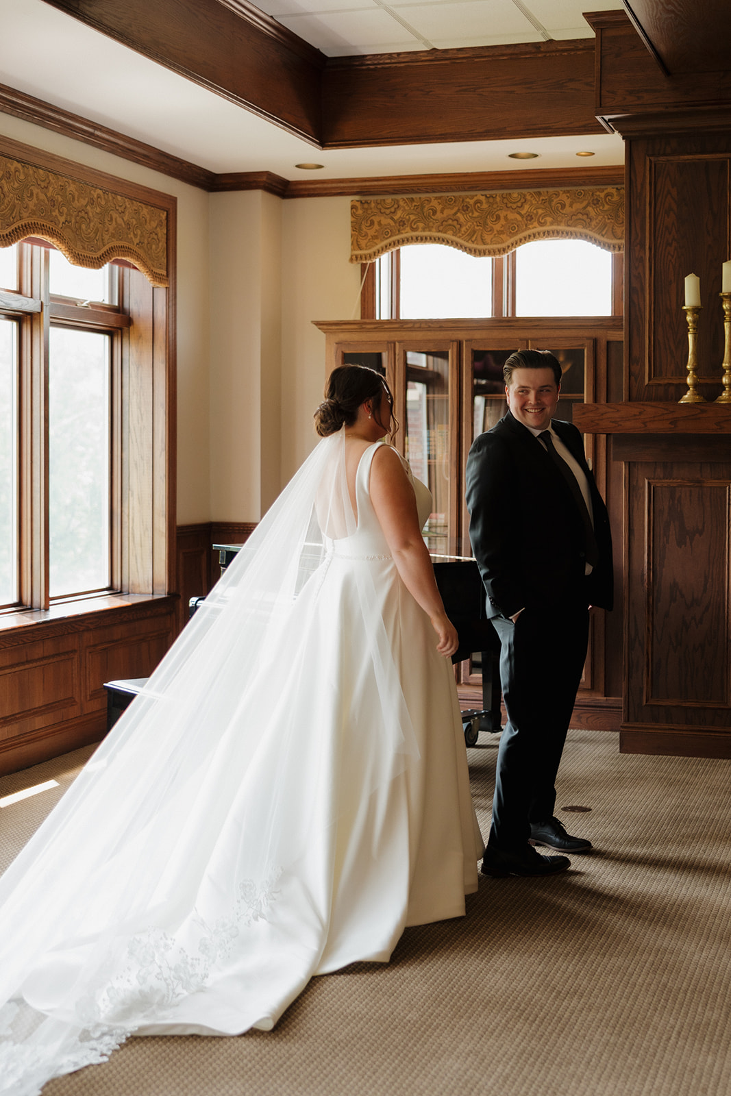 Bride and groom smiling at each other during their first look at a country club wedding, with her long veil trailing behind her.