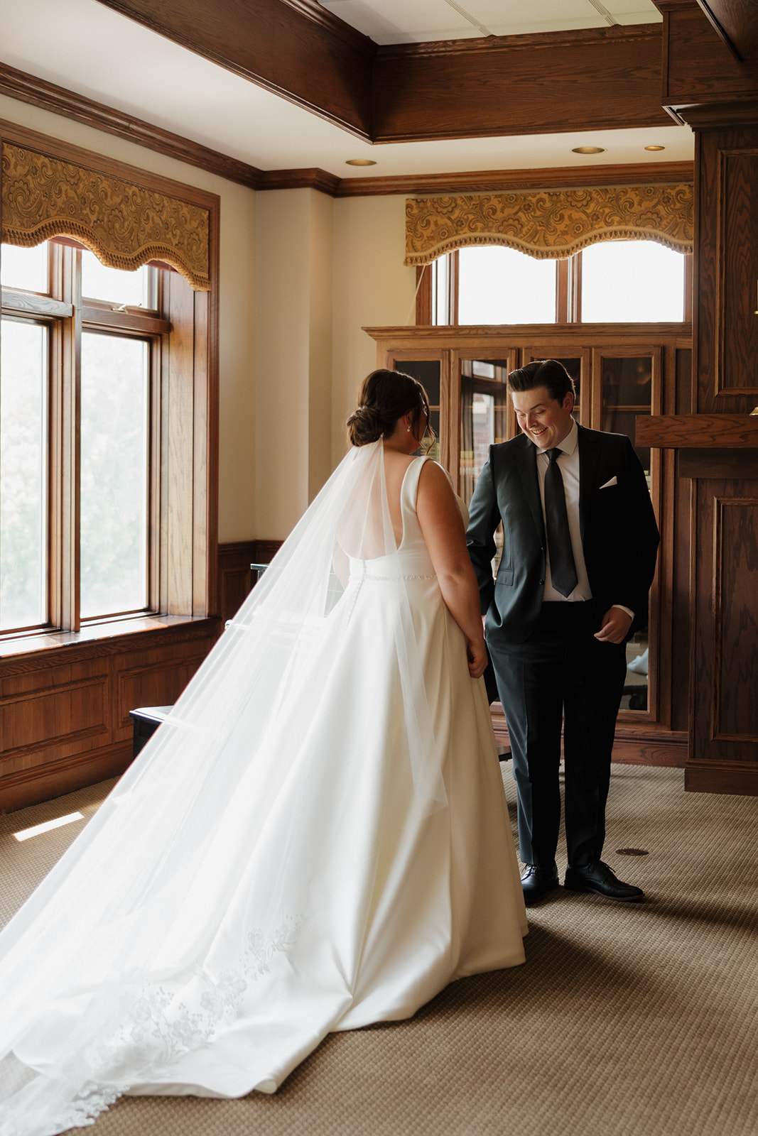 Groom seeing the bride for the first time during an emotional first look at their country club wedding, both standing in a library-style room with wood-paneled walls.