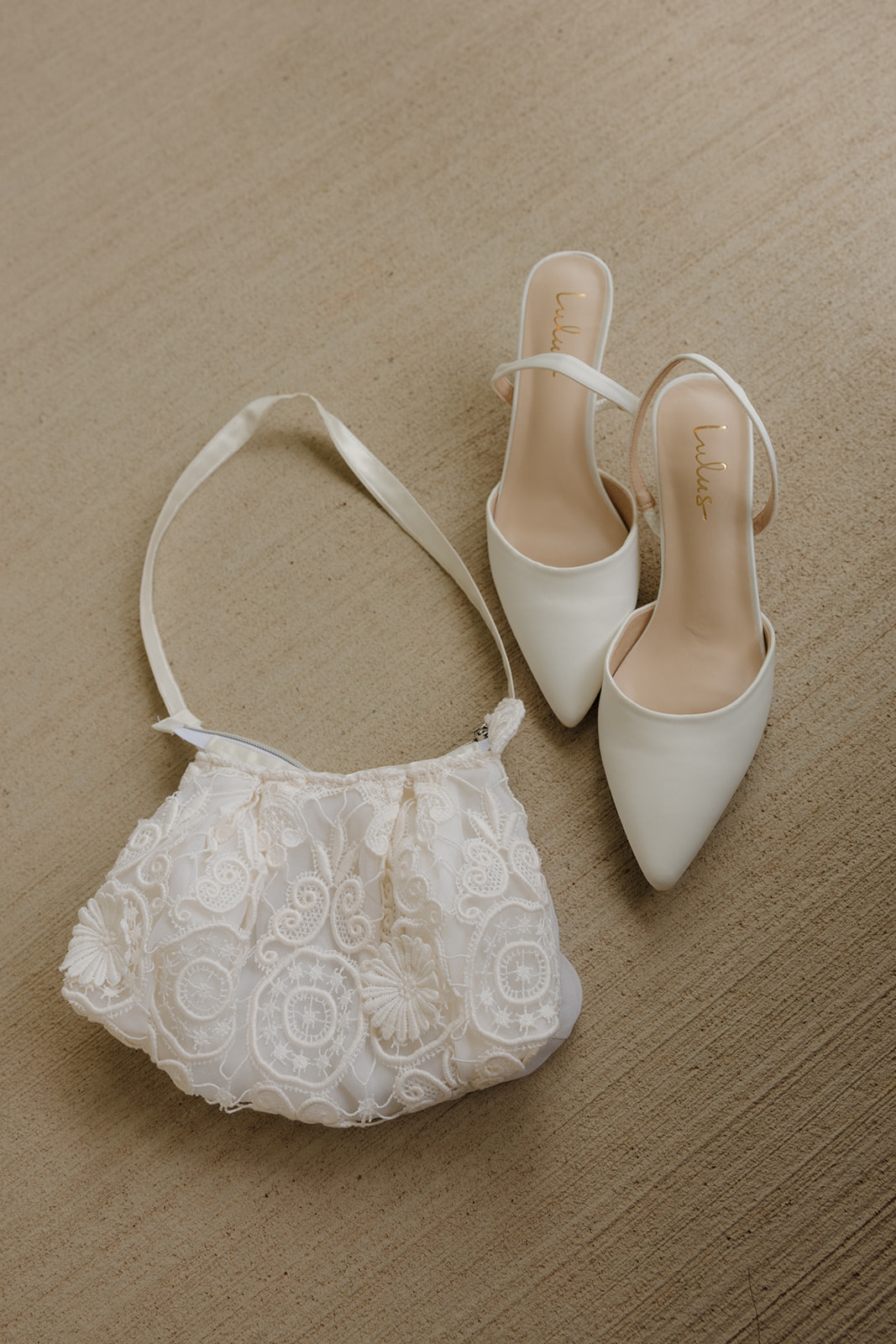 Creamy white pointed-toe heels and a delicate lace clutch bag laid out on neutral-toned carpet for a classic country club wedding flat lay.