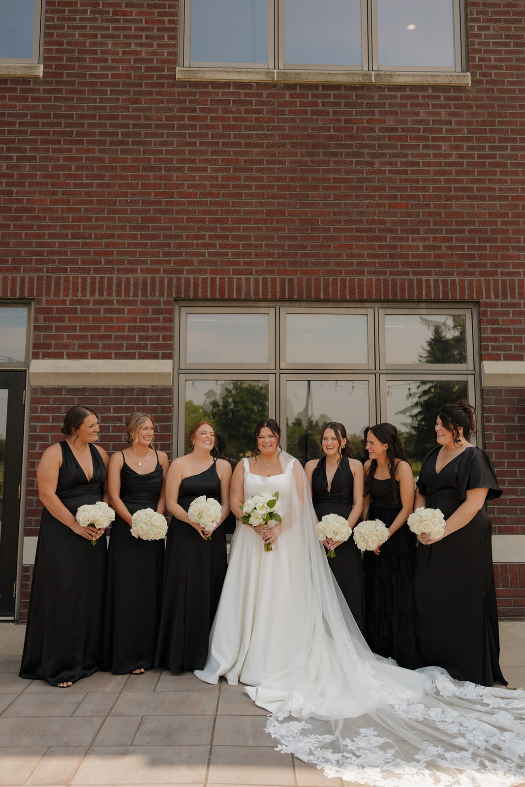 Bride standing with her bridesmaids, all in black dresses holding white bouquets, in front of the brick exterior at a country club wedding.