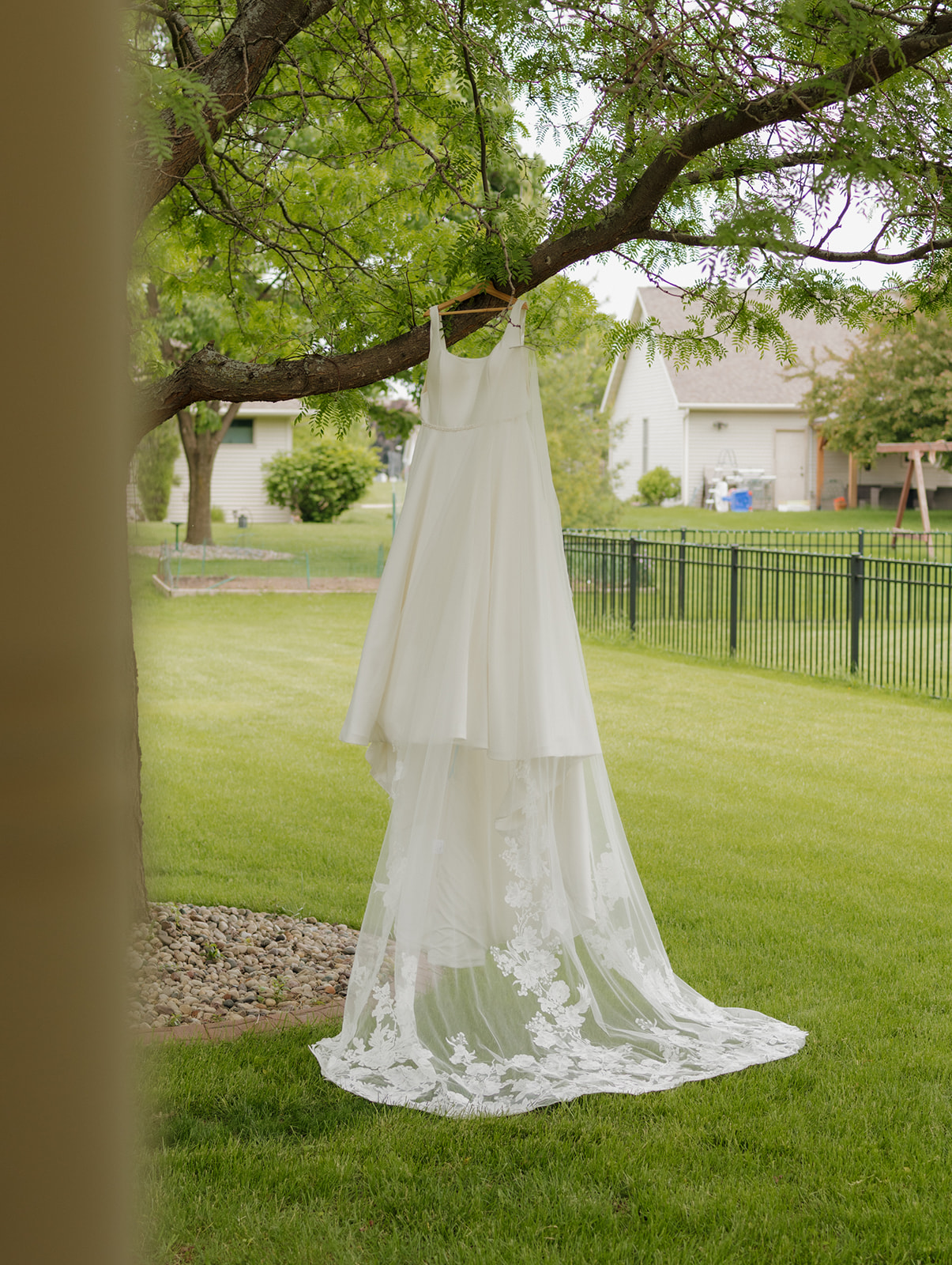 Bride’s wedding dress with a long lace train hanging from a tree branch outside the country club venue, ready for a summer wedding day.
