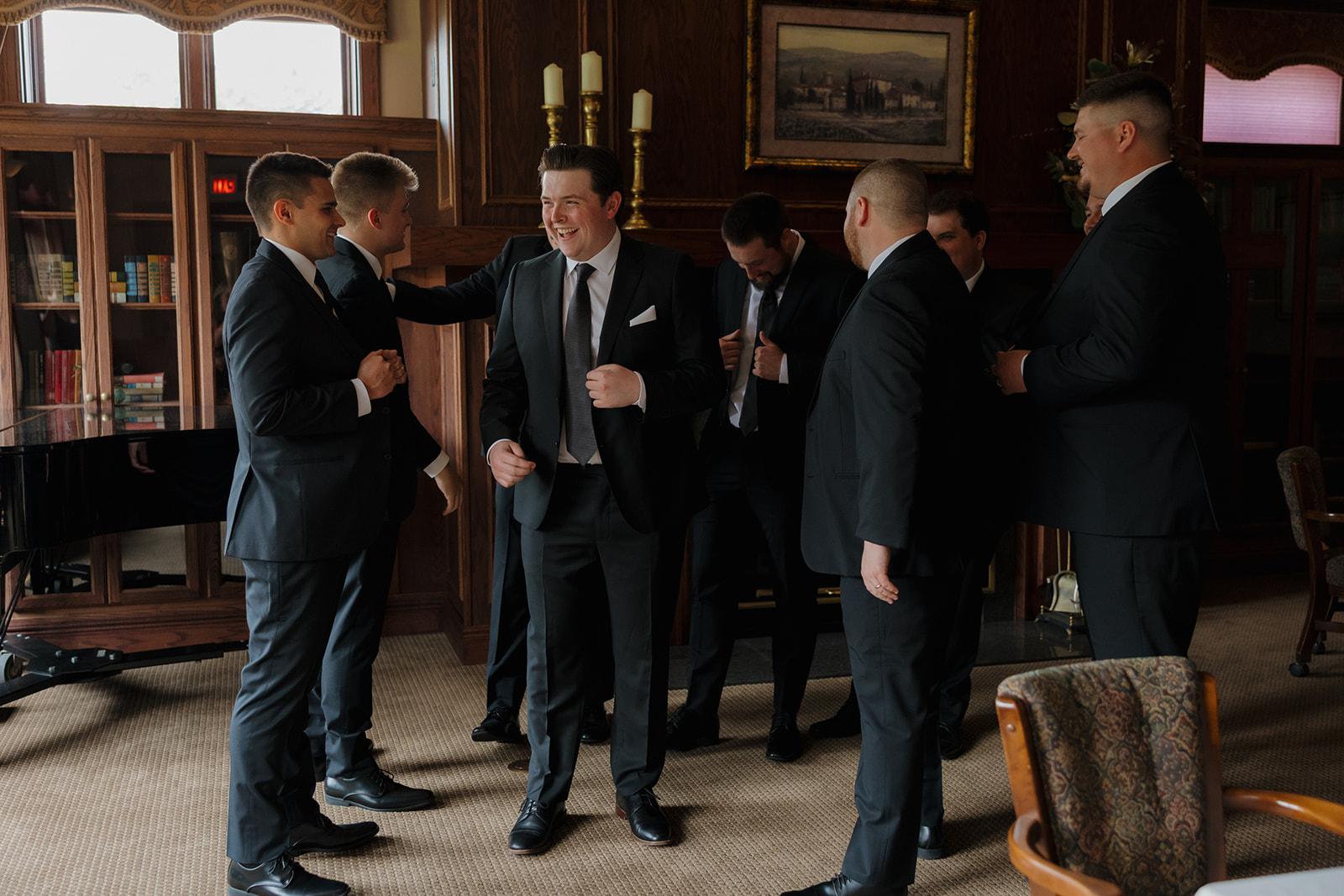 Groom laughing with his groomsmen in a wood-paneled library lounge at a country club wedding, all dressed in classic black suits.