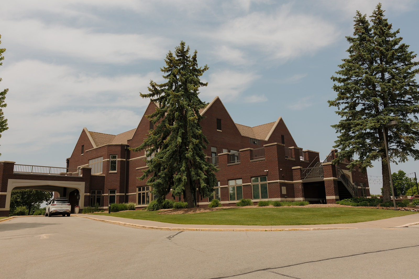 Wide shot of a red-brick country club venue with classic architecture, tall trees, and a covered drive, captured on a sunny wedding day.