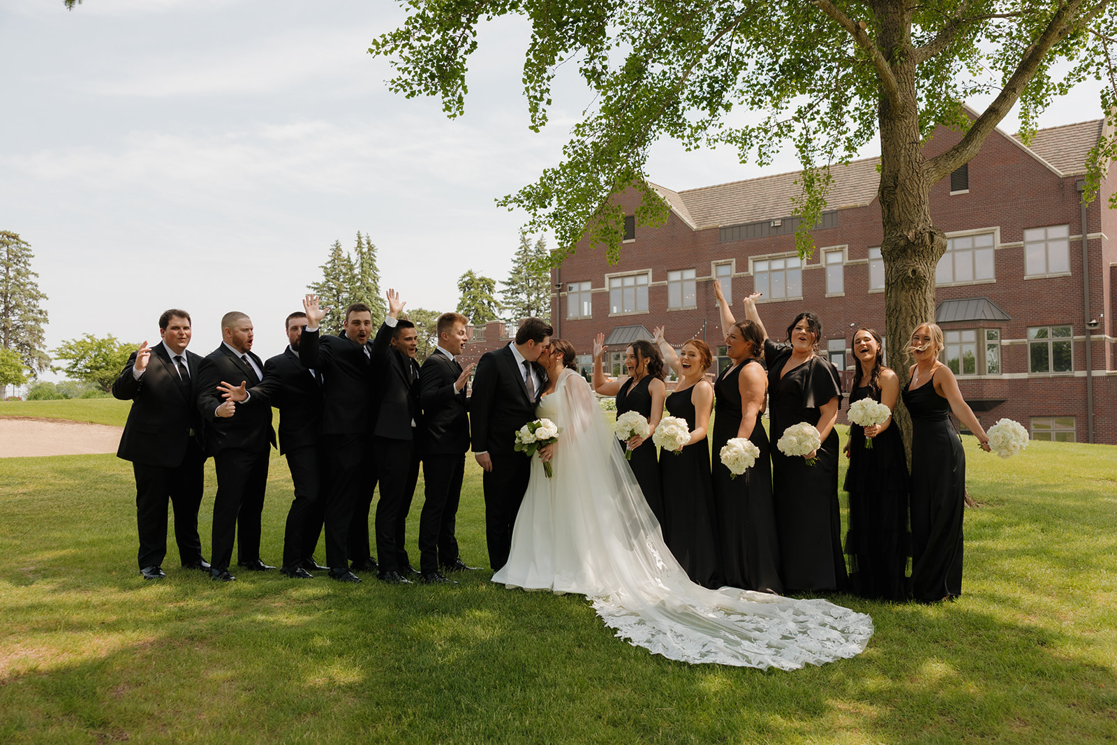Bride and groom share a kiss as the wedding party cheers around them on the lawn of a brick country club wedding venue.