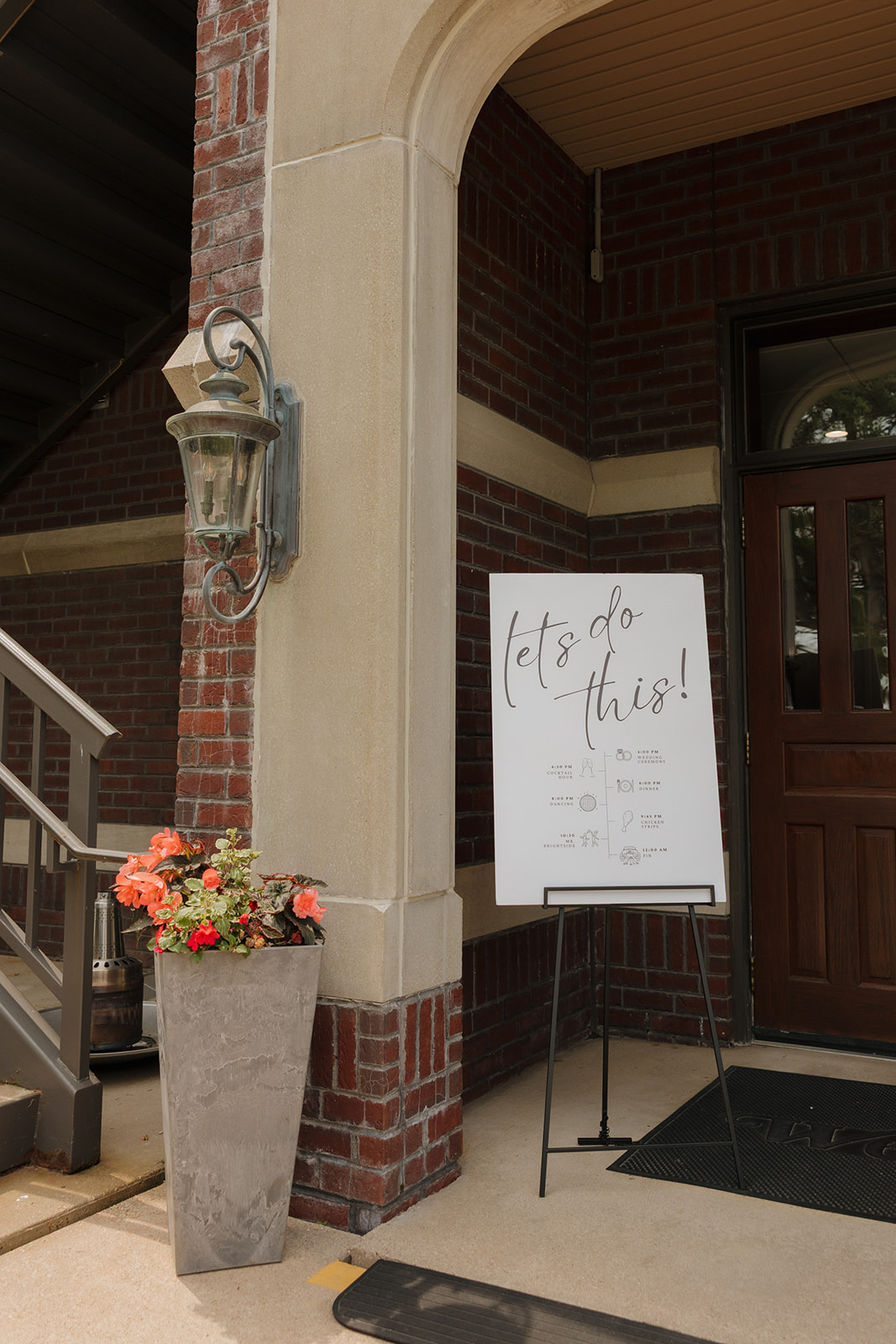 Modern wedding welcome sign reading “Let’s do this!” displayed outside a brick country club wedding venue, with a tall floral planter and vintage lantern nearby.