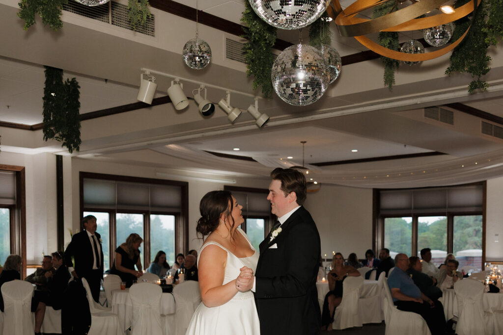 Bride and groom sharing their first dance under hanging greenery and disco balls during their country club wedding reception.