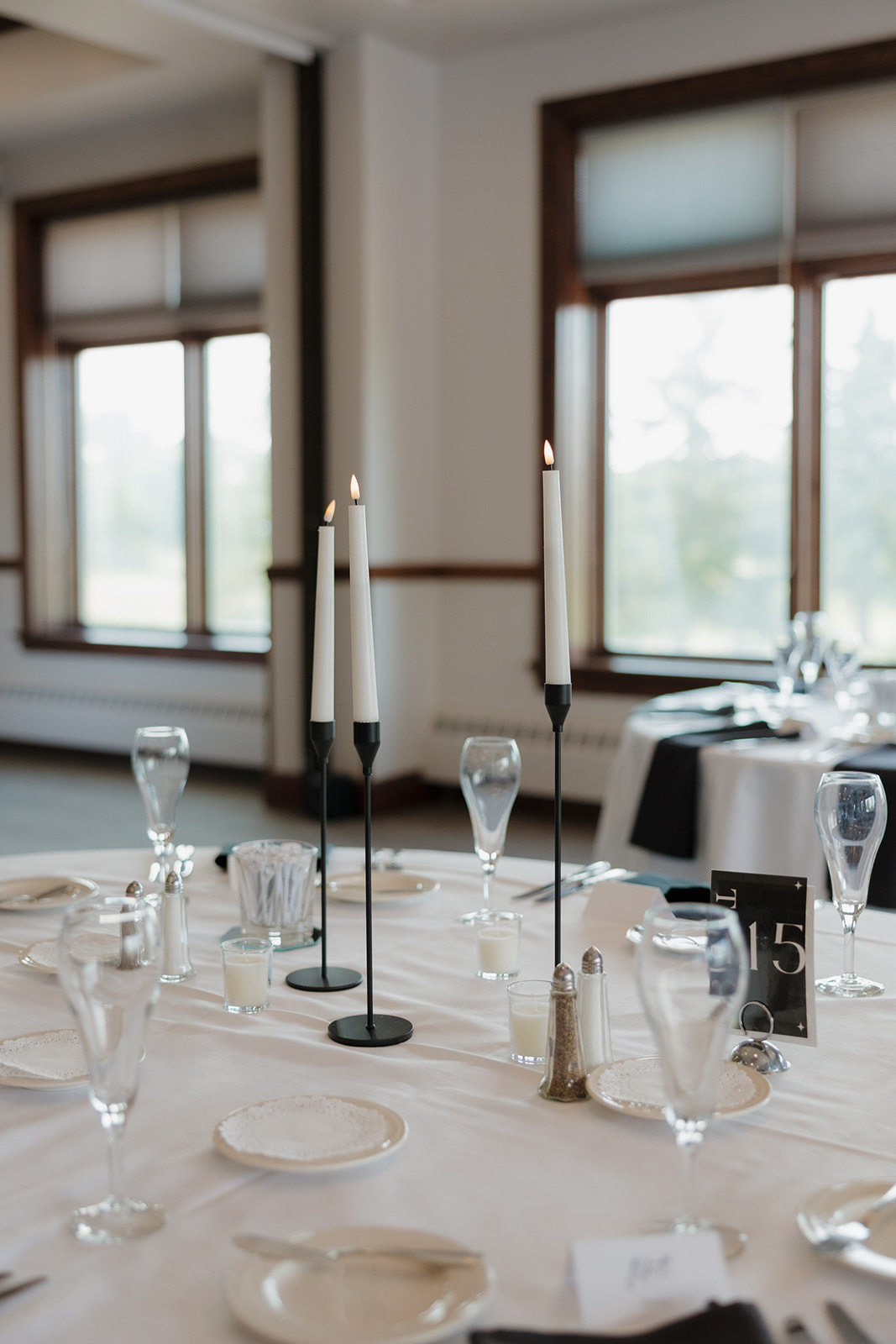 Minimalist country club wedding reception table with tall black candle holders, champagne flutes, and table number 15 in front of large sunlit windows.