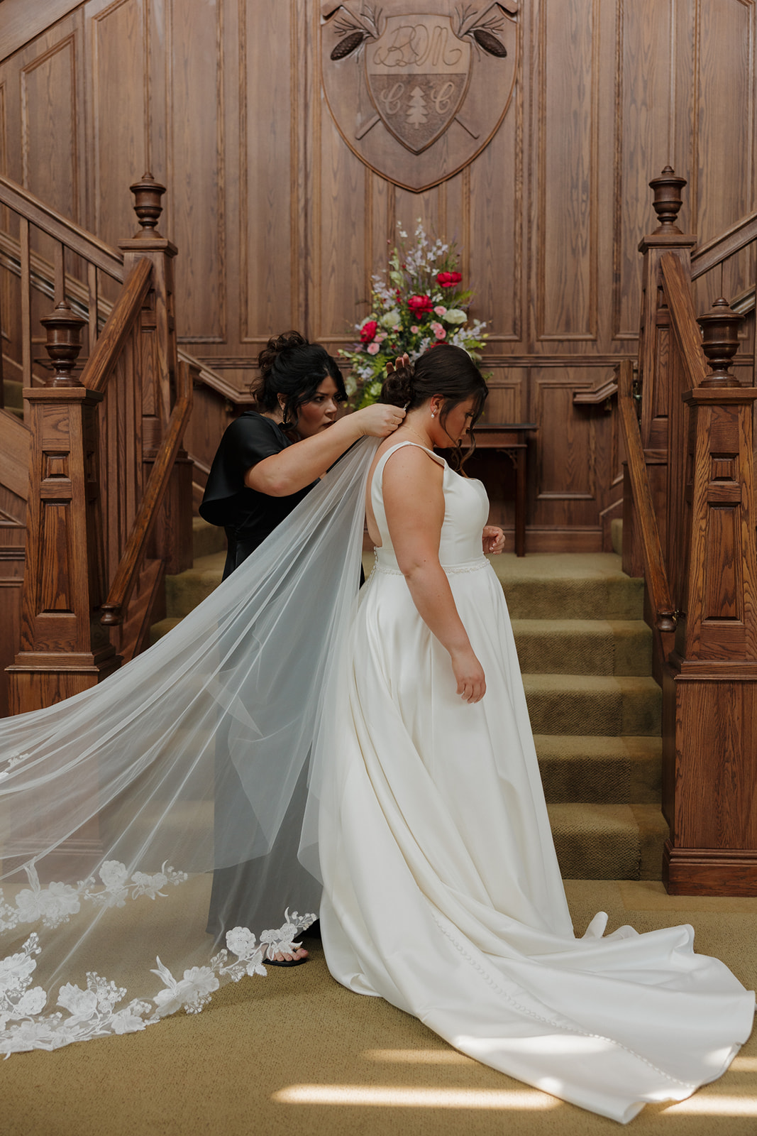 Bride getting her veil secured by a bridesmaid on the grand staircase of a country club wedding venue, with wood paneling and vibrant florals in the background.