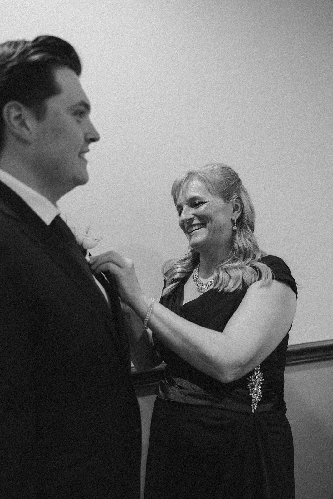 Black and white candid of the groom smiling as his mother pins his boutonnière, sharing a sweet pre-ceremony moment at their country club wedding.