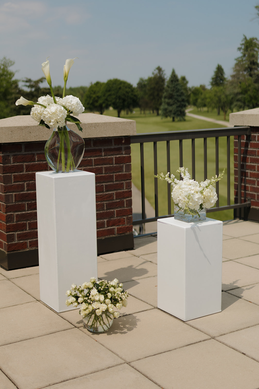 White floral arrangements displayed on modern pedestals overlooking the golf course at a country club wedding ceremony setup.