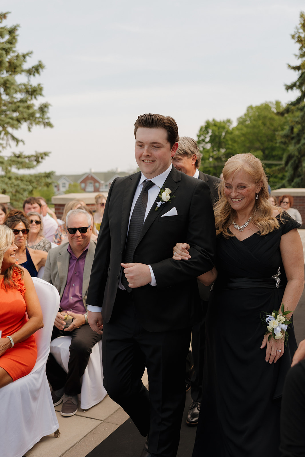 Groom walking arm-in-arm with his mom down the aisle before the ceremony begins, surrounded by seated guests at a country club wedding.