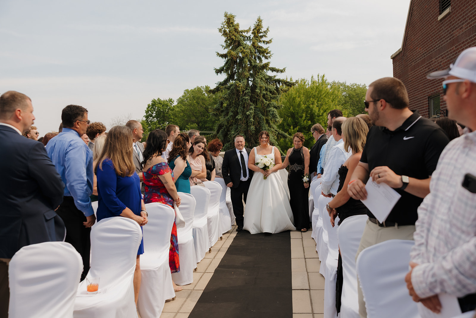 Bride walking down the aisle with her parents at an outdoor country club wedding ceremony as guests stand and watch.