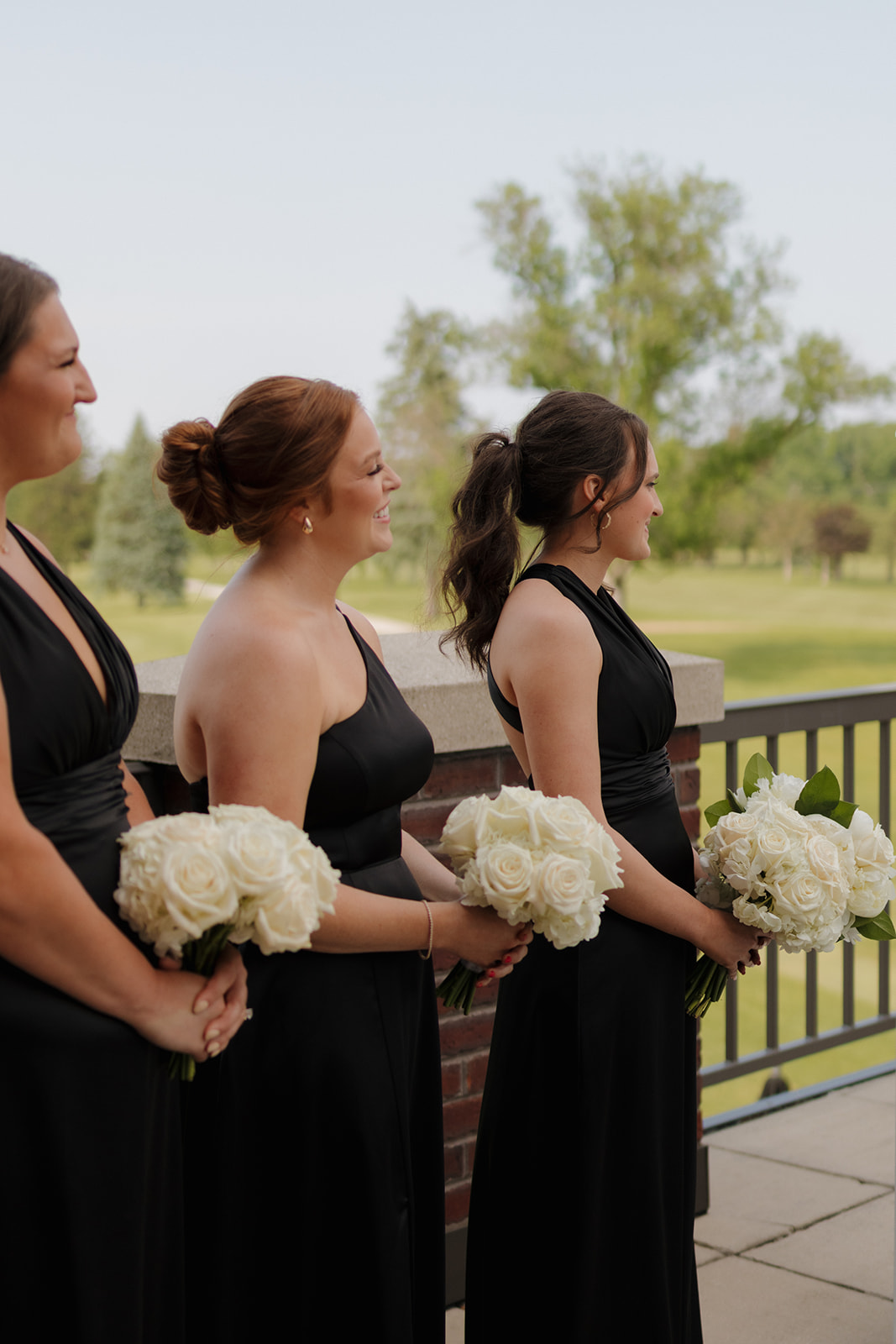 Bridesmaids smiling and holding white rose bouquets during the ceremony at a summer country club wedding, with the golf course in the background.