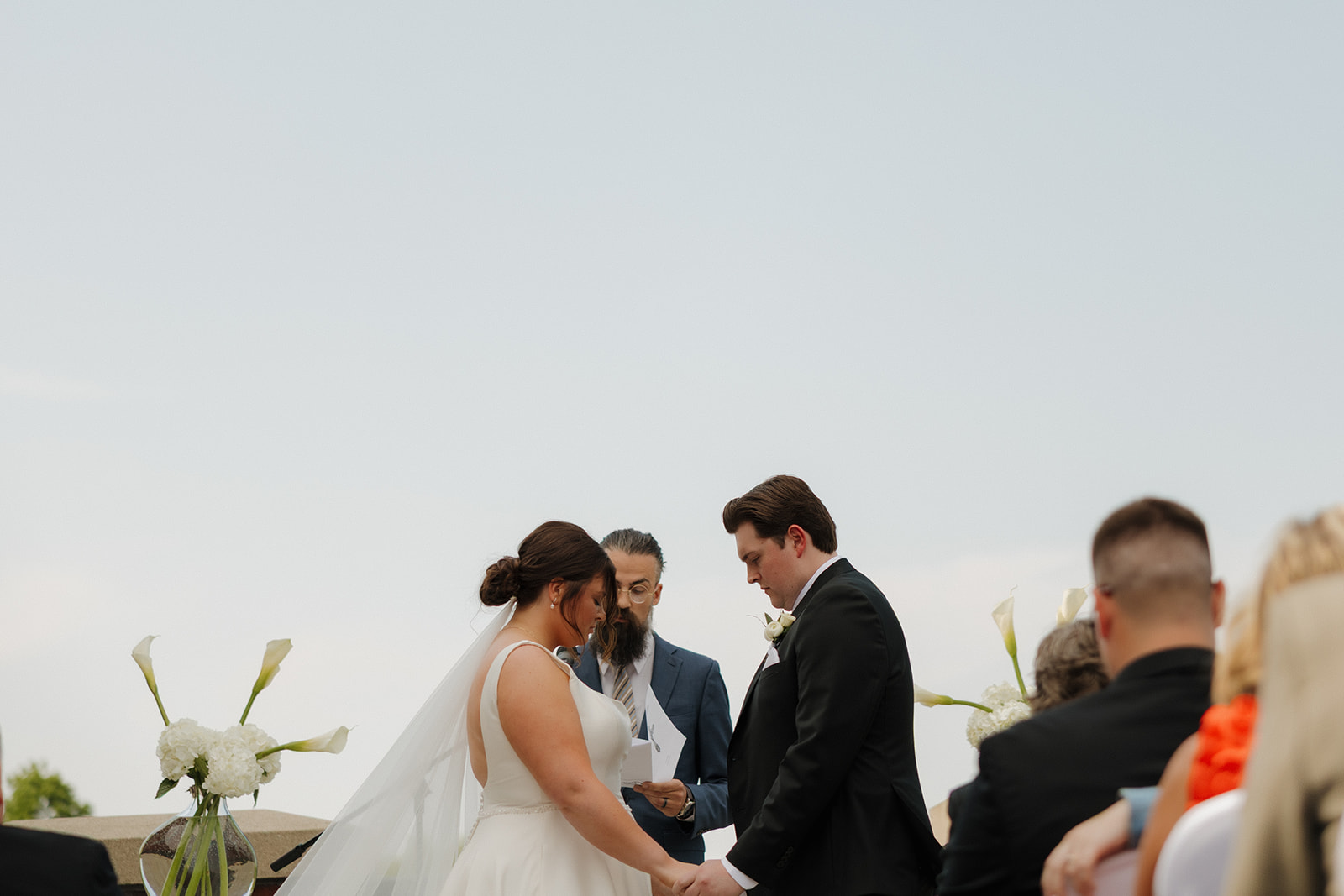 Bride and groom holding hands and bowing their heads during a heartfelt moment in their country club wedding ceremony, with white floral arrangements on either side.