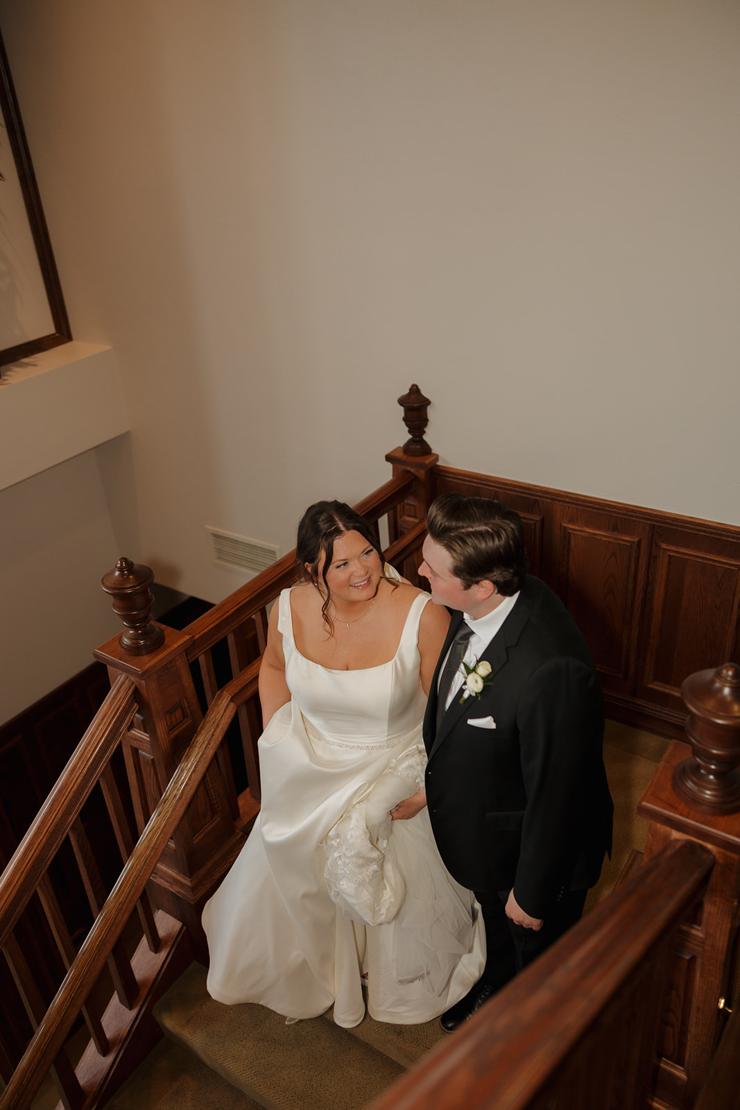 Bride and groom smiling at each other while walking up the wood-paneled staircase of their country club wedding venue.