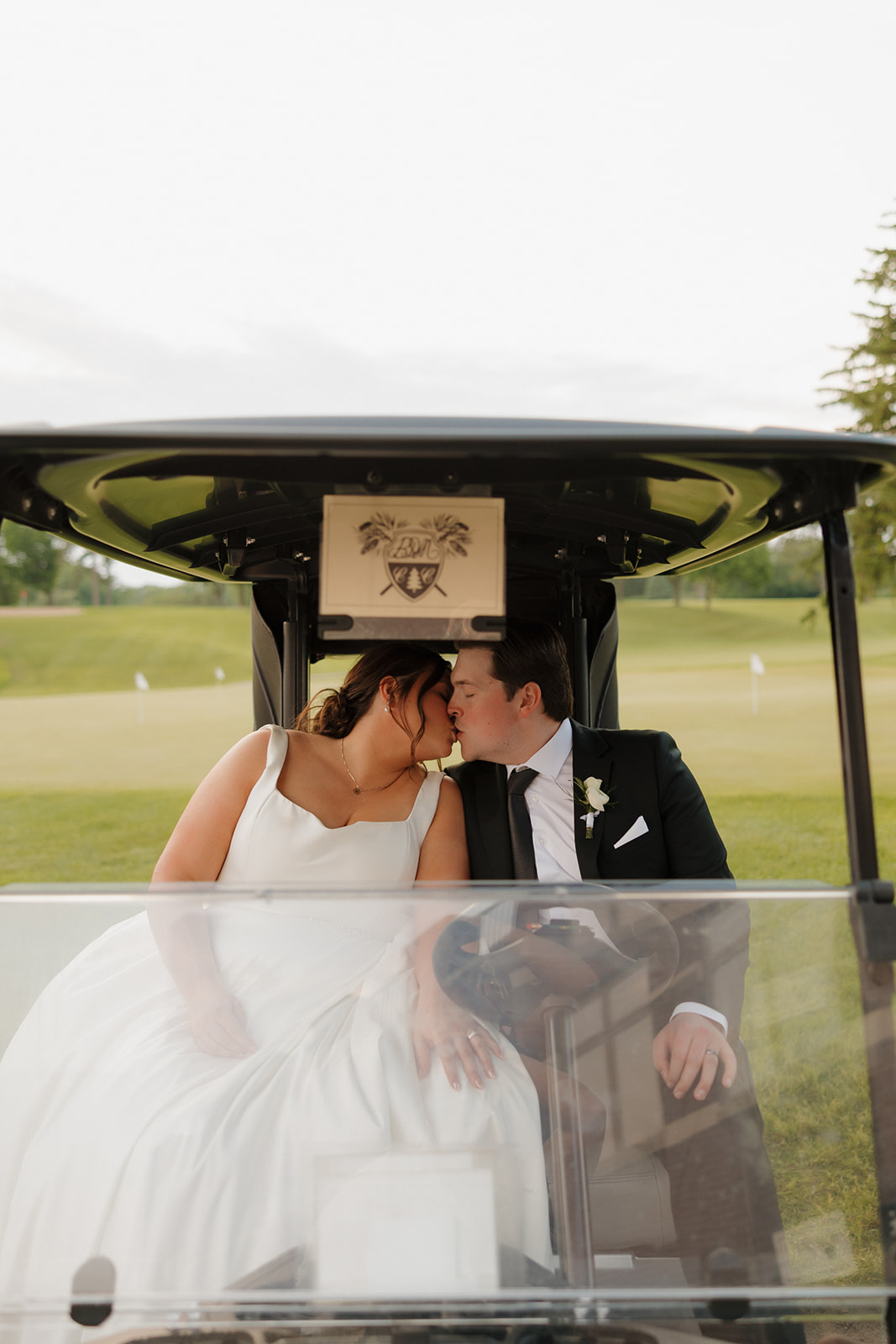 Bride and groom share a kiss inside a golf cart on the course at their country club wedding, framed by manicured greens and soft sunset light.
