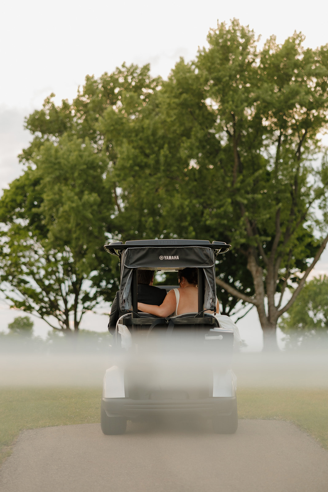 Bride and groom captured from behind as they ride away in a Yamaha golf cart after their country club wedding ceremony, surrounded by trees and golden light.
