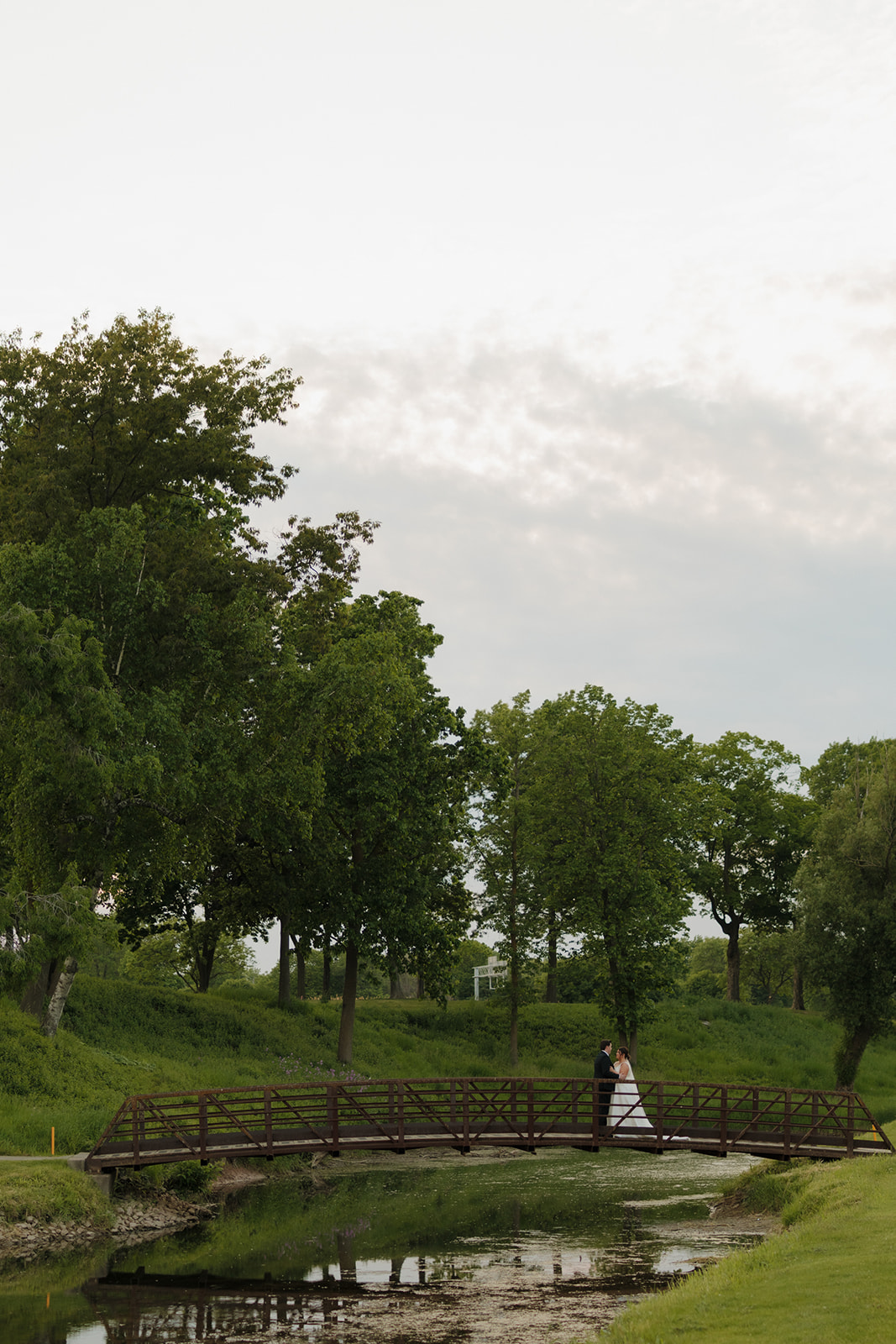 Bride and groom kissing on a wooden bridge over a pond, surrounded by trees on the lush golf course at their country club wedding.