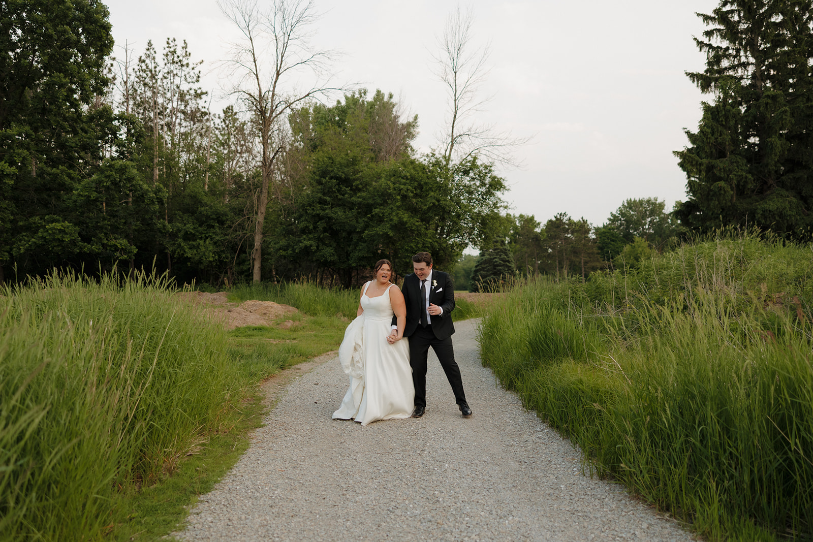 Joyful bride and groom walking hand-in-hand down a gravel path surrounded by tall grass and trees during their country club wedding portraits.